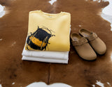 Folded yellow shirt with black design and white pants on a brown cowhide rug, accompanied by beige clogs.