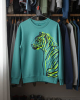 Teal sweatshirt with a tiger design hanging on a rack among other clothes. fauna kids